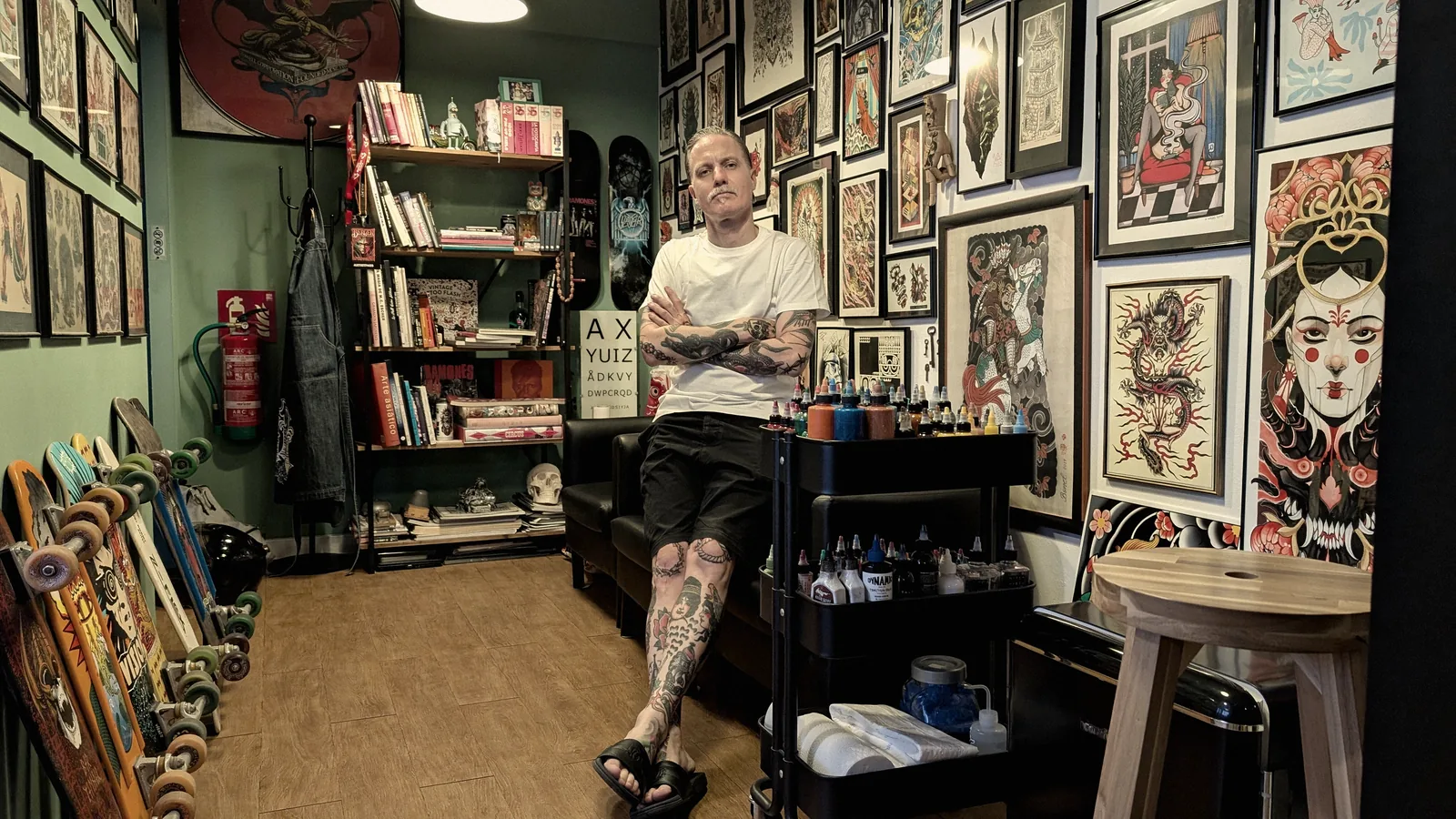 Sala principal de A Sailor's Grave Tattoo en Granollers: paredes de láminas enmarcadas, estantería de libros de tatuaje, monopatines apoyados y el carro de tintas junto a la camilla.