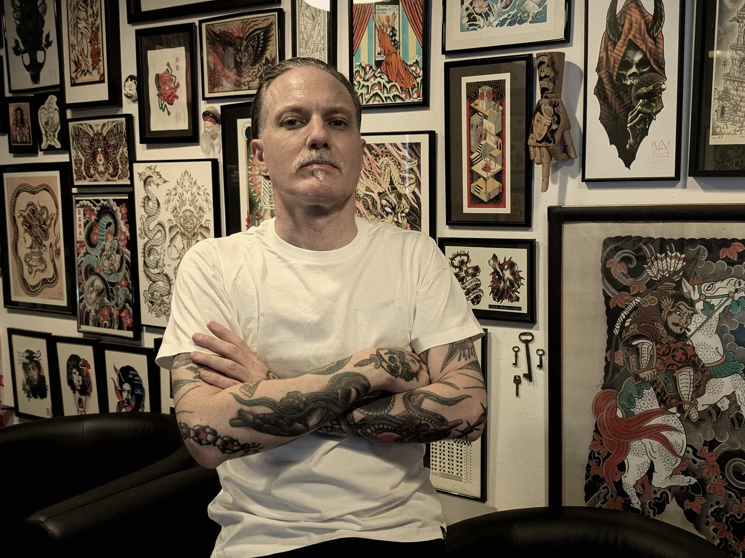 Leo Martínez with arms crossed at A Sailor's Grave Tattoo in Granollers, in front of the studio's wall of framed flash.
