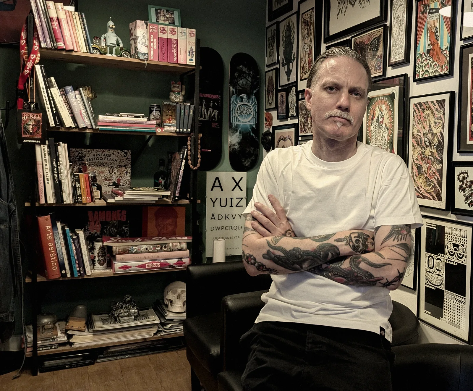 Leo Martínez de brazos cruzados en A Sailor's Grave Tattoo, su estudio privado de Granollers, con la estantería de libros de tatuaje y la pared de láminas enmarcadas.