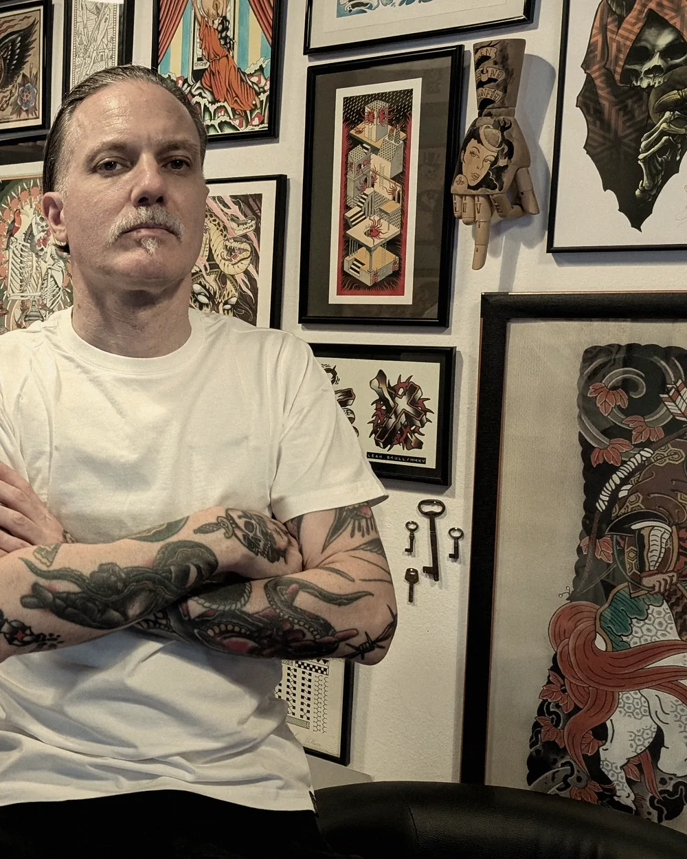 Leo Martínez seated in the main room at A Sailor's Grave Tattoo, Granollers, with the wall of framed flash behind him.